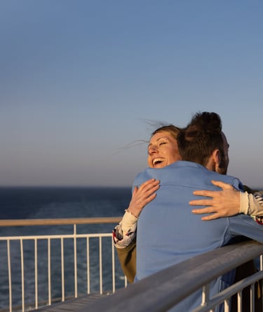 To personer som omfavner hverandre på en balkong med utsikt over havet.