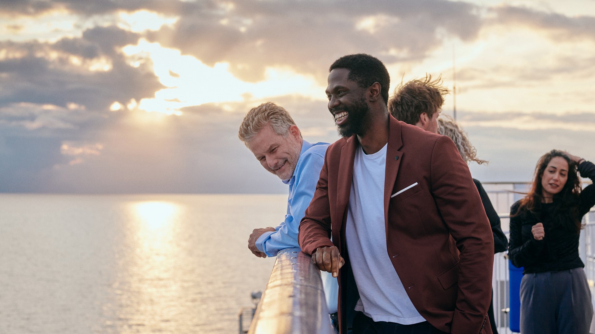 Fire personer som står på et dekk med havet og solnedgang i bakgrunnen.