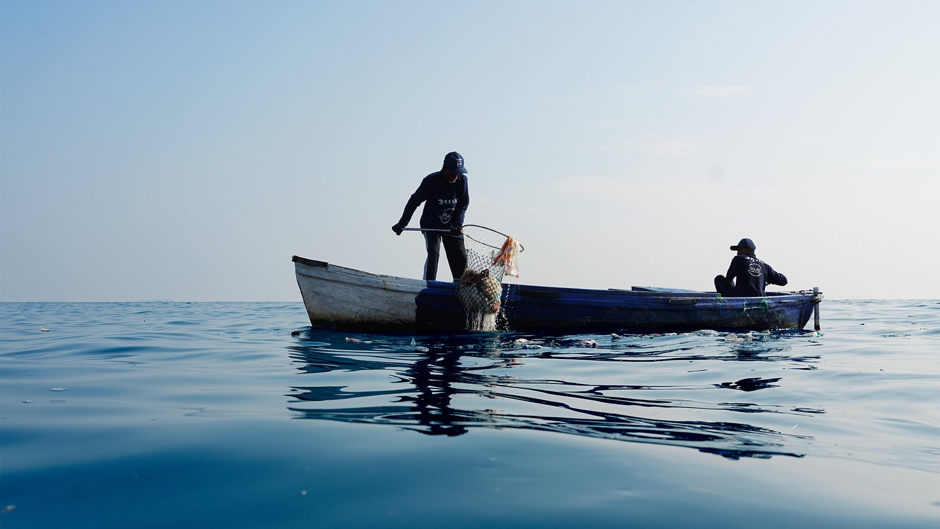 To personer på en liten båt midt på et rolig vann, hvor en håndterer et fiskegarn.
