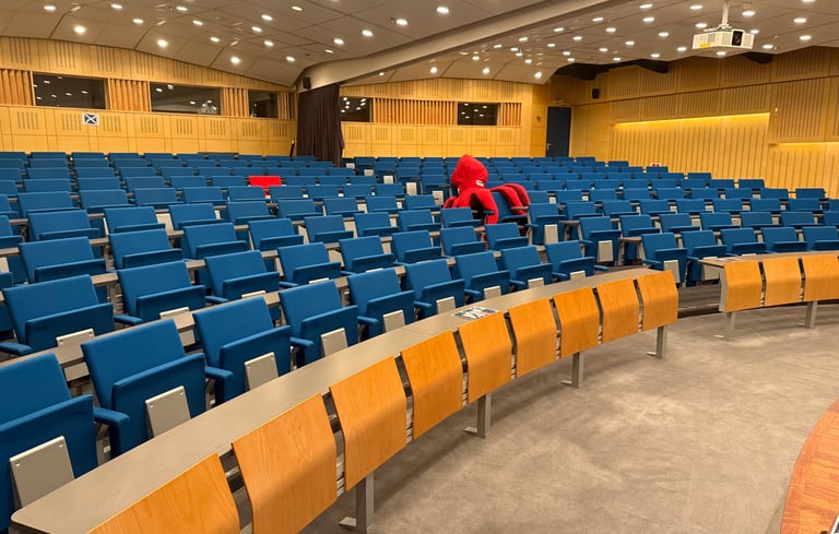 Tomt auditorium med blå stoler og trepulter, én person i rødt sitter alene