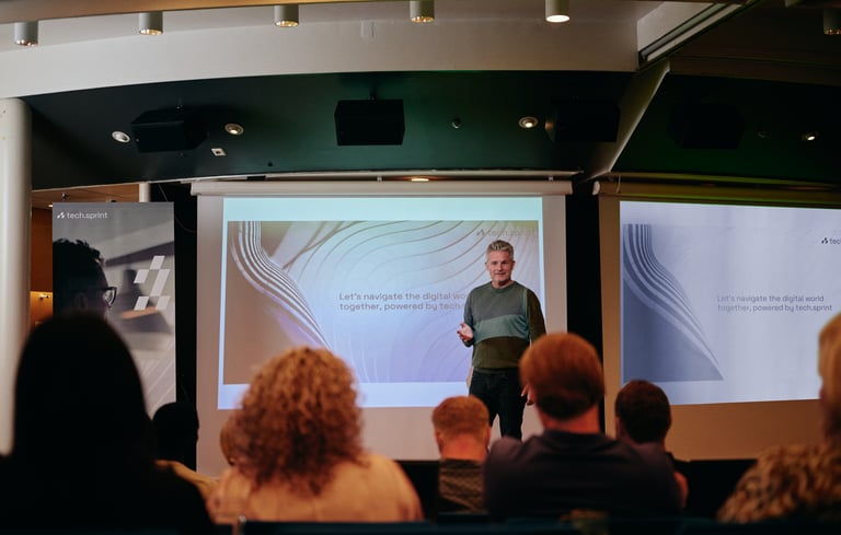 Person som holder en presentasjon foran en skjerm med publikum sittende.